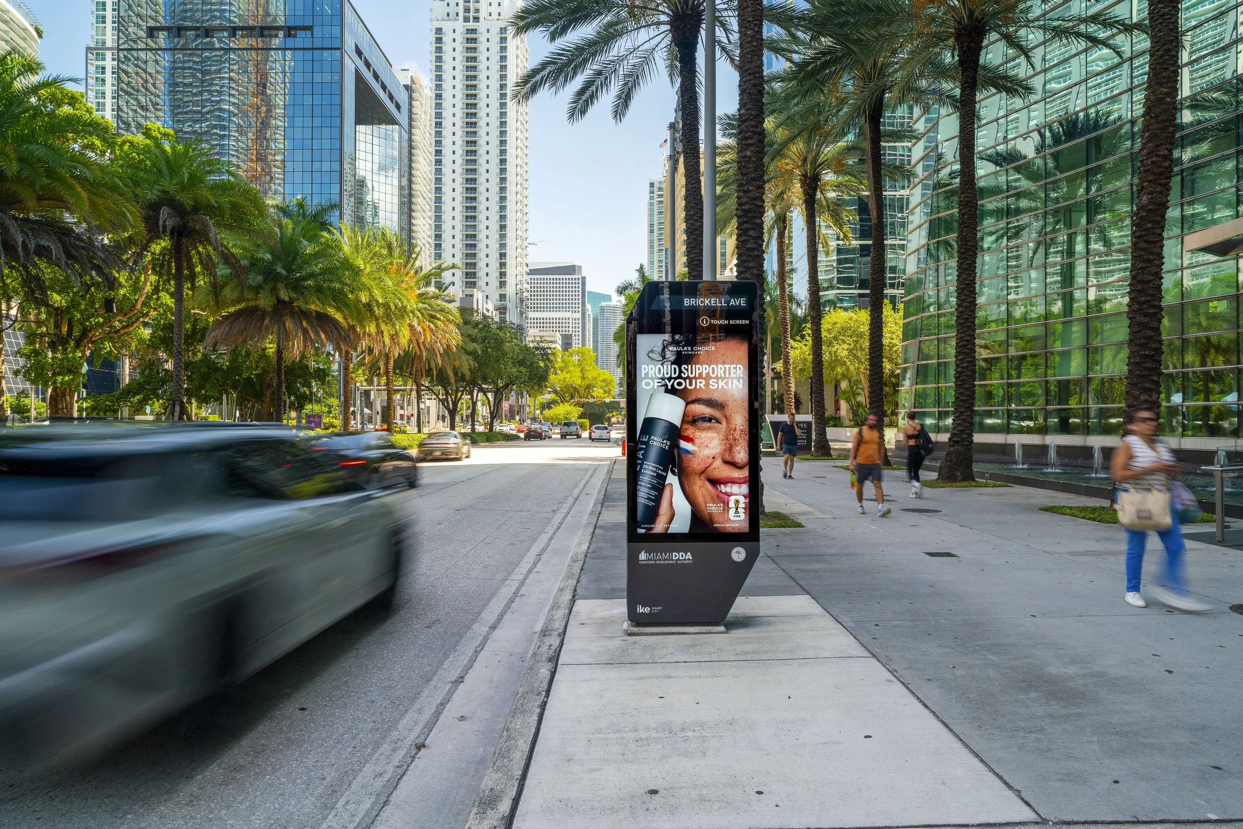 City street scene near Miami, Florida, with tall glass skyscrapers, palm trees, and a digital billboard advertising skincare products. Cars are moving along the street, and pedestrians walk on the sidewalk.