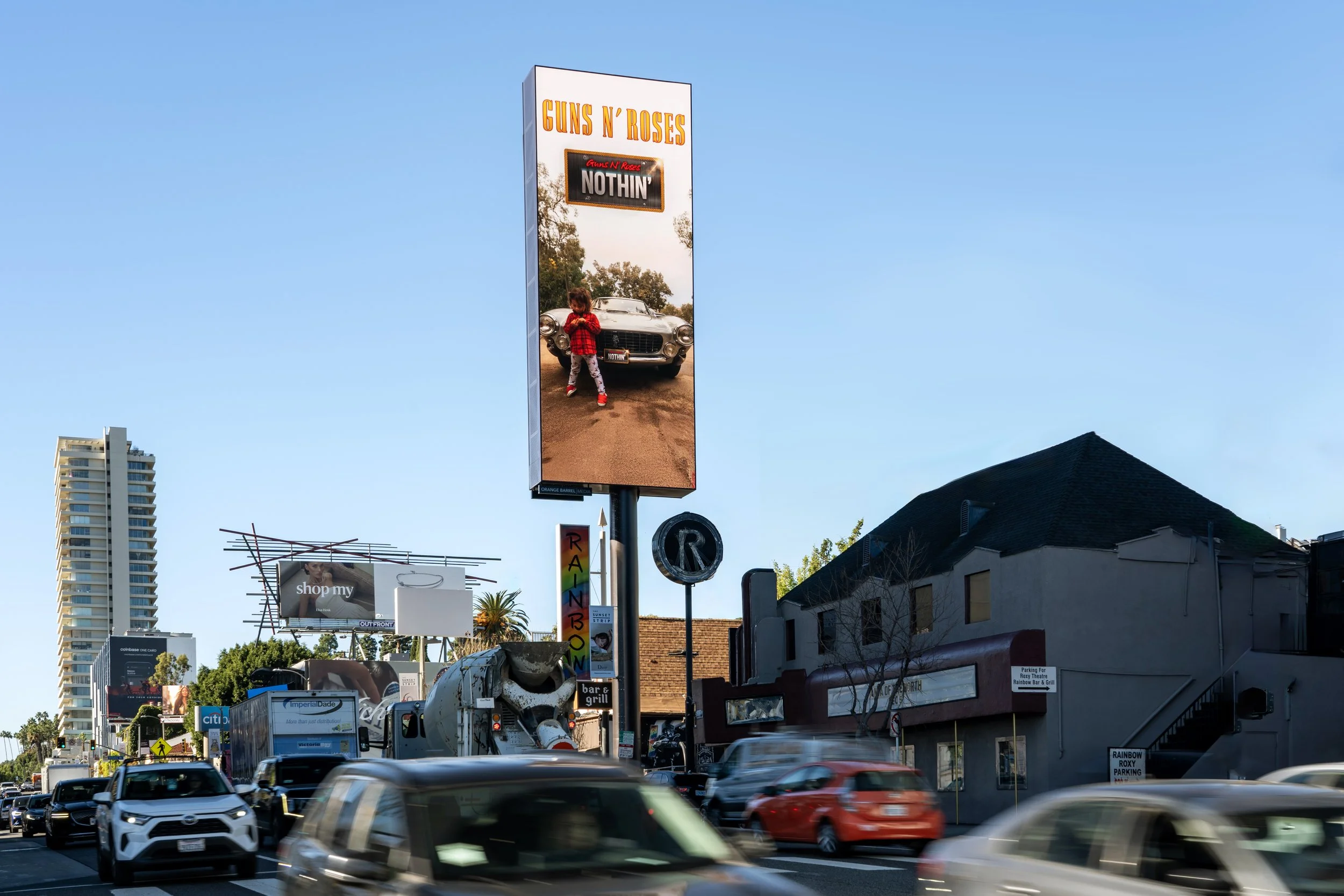 A busy city street with cars and trucks, billboard advertising Guns N' Roses, and various storefronts, including a rainbow-colored bar and grill.