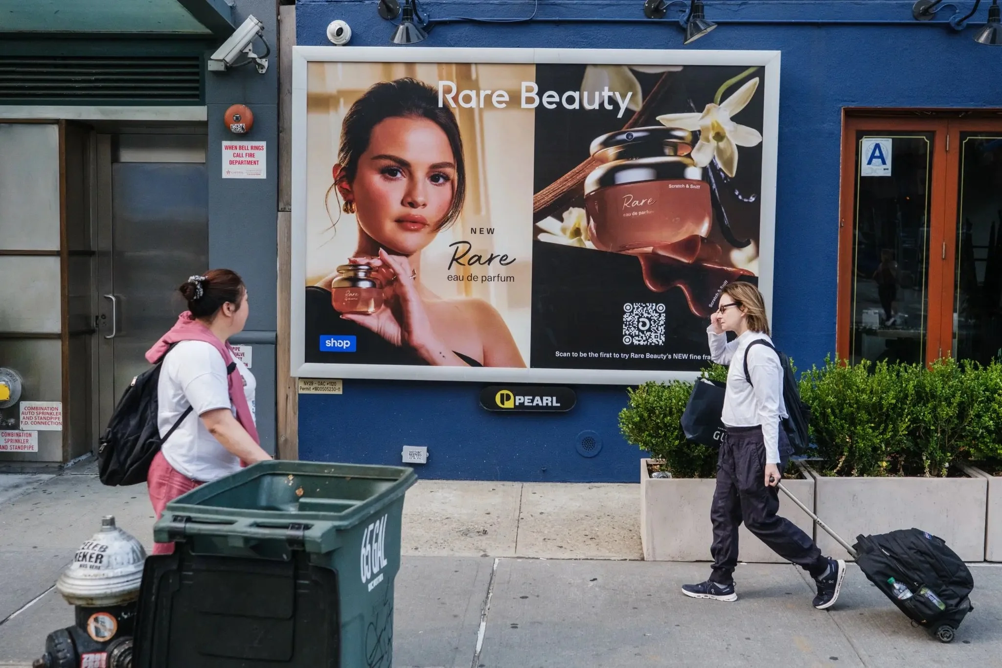 Two women walking past a large advertisement for Rare Beauty perfume on a blue wall. The woman on the left has a backpack and is wearing a pink scarf over a white shirt. The woman on the right is wearing glasses, a white jacket, black pants, and is pulling a rolling suitcase. There is a green trash bin and a fire hydrant on the sidewalk.