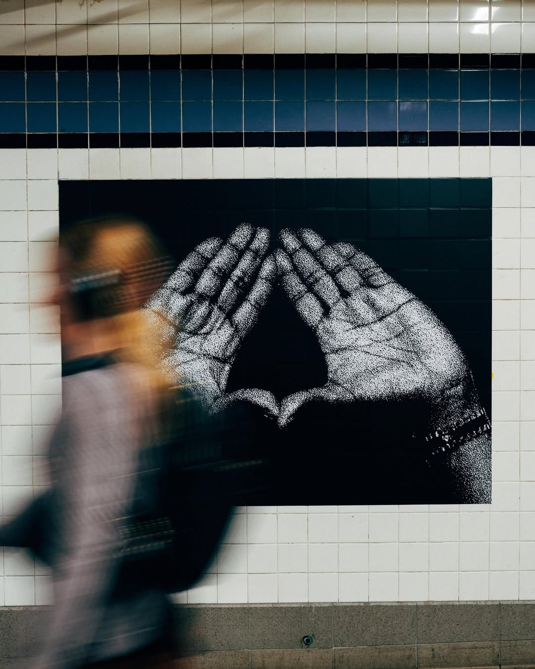 A person walking past a black and white mural of hands forming a diamond shape on a tiled wall, with the person appearing blurred in motion.