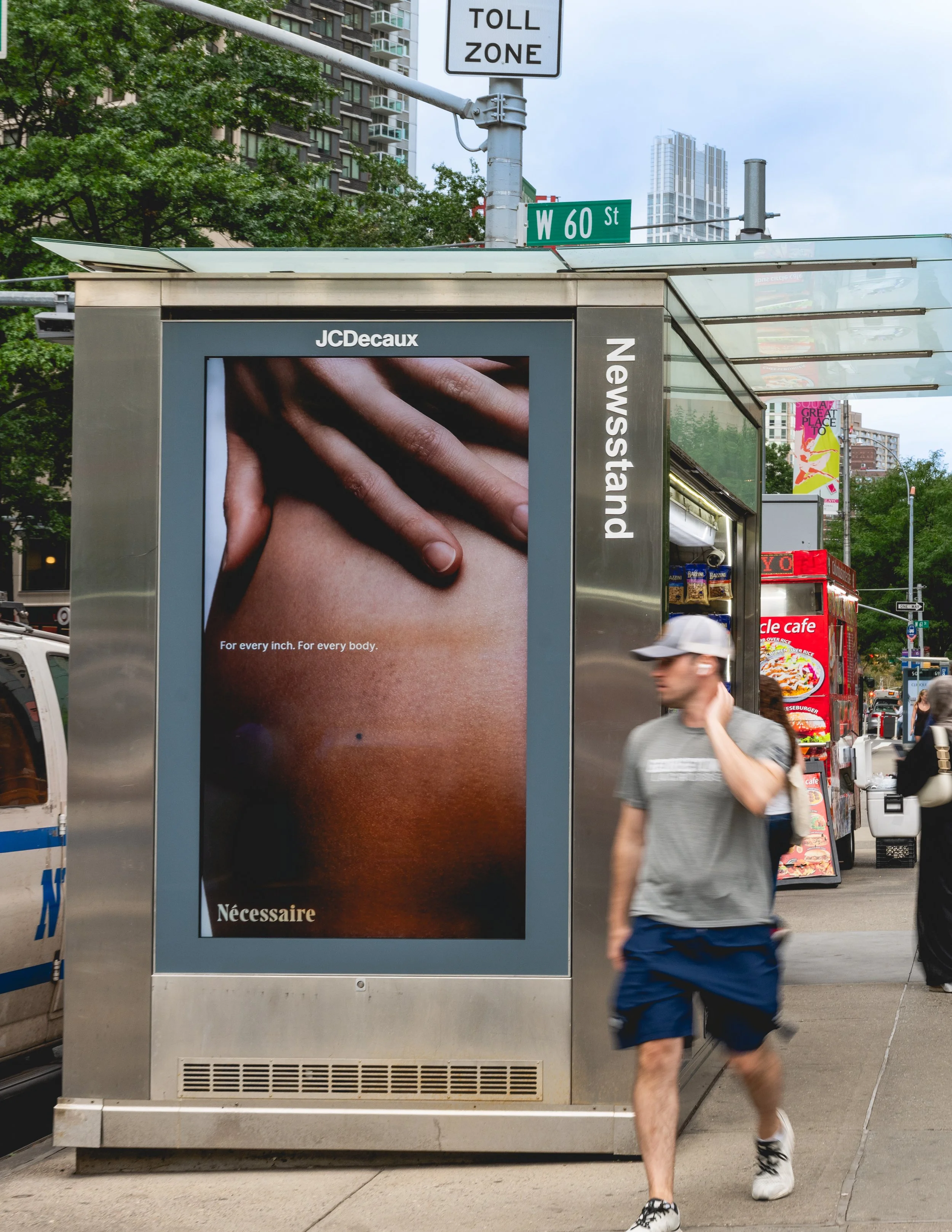 A street scene featuring a bus shelter with an advertisement showing a close-up of a hand on a person's body and a caption reading 'For every inch. For every body.' The bus shelter is located at the intersection of West 60th Street, near a city high-