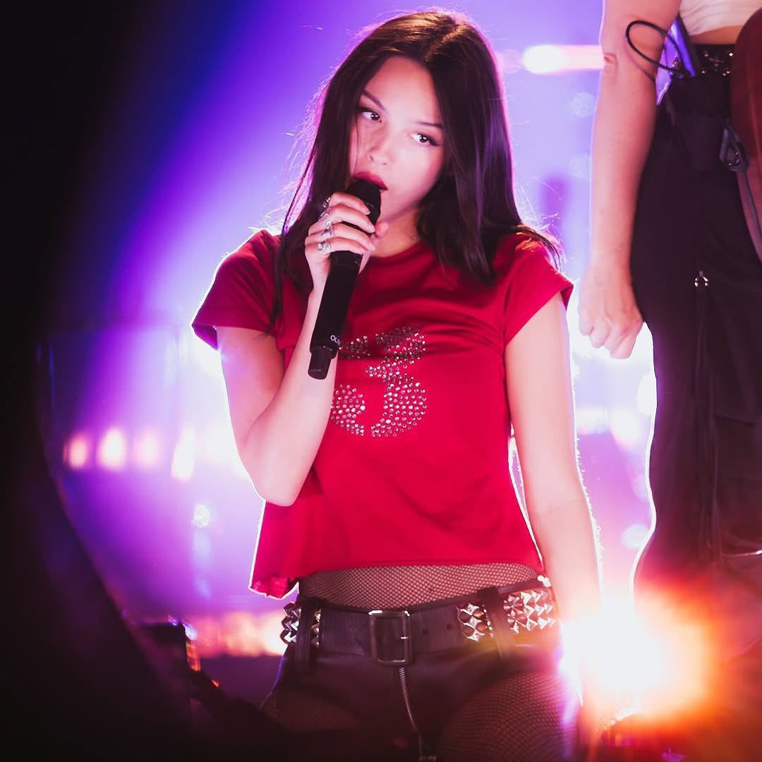 Olivia Rodrigo with dark hair singing into a microphone on stage, wearing a red shirt with a sparkling design, black shorts with studs, and fishnet stockings, with colorful stage lighting behind her.