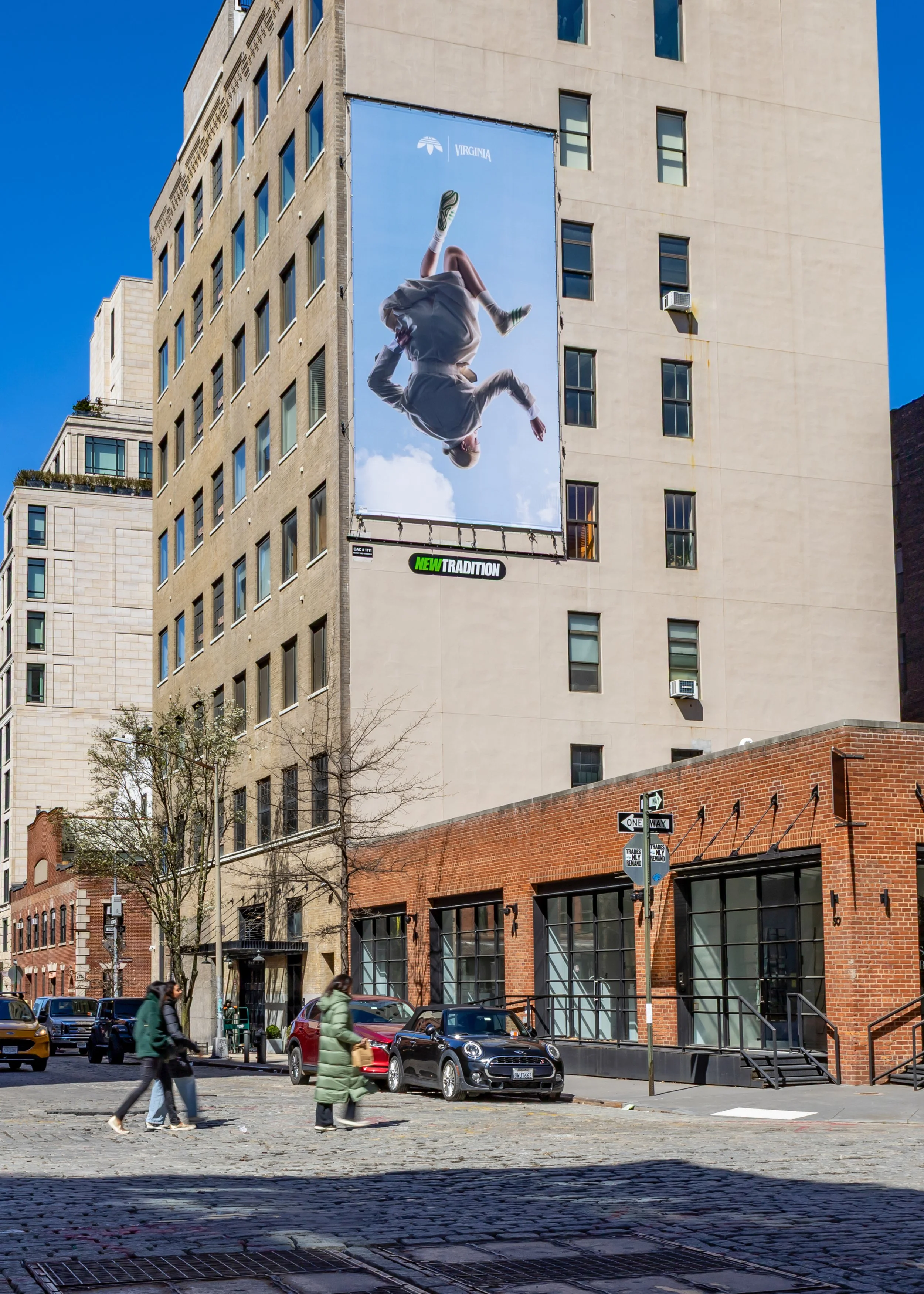 City street scene with people crossing, parked cars, and a large building featuring an Adidas billboard with a diver and the words 'New Tradition'.
