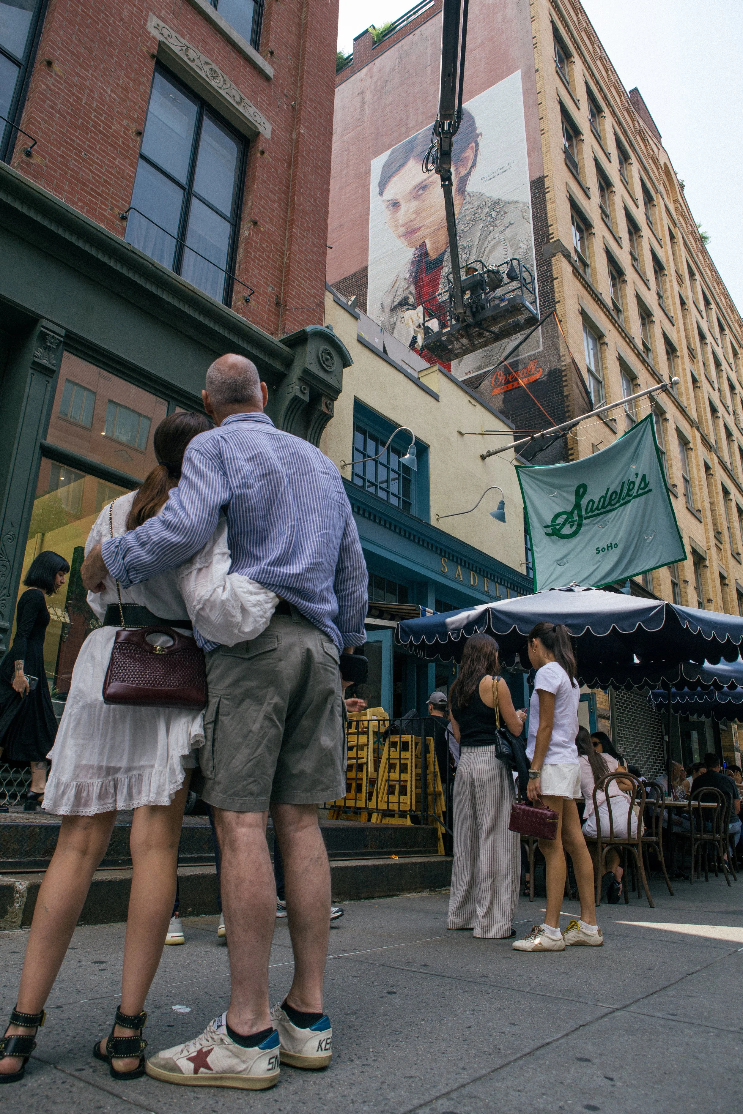 People standing and talking outside in front of a building with a large mural of Gracie Abram's face on it. The scene appears to be in an urban area, possibly a city street or plaza.