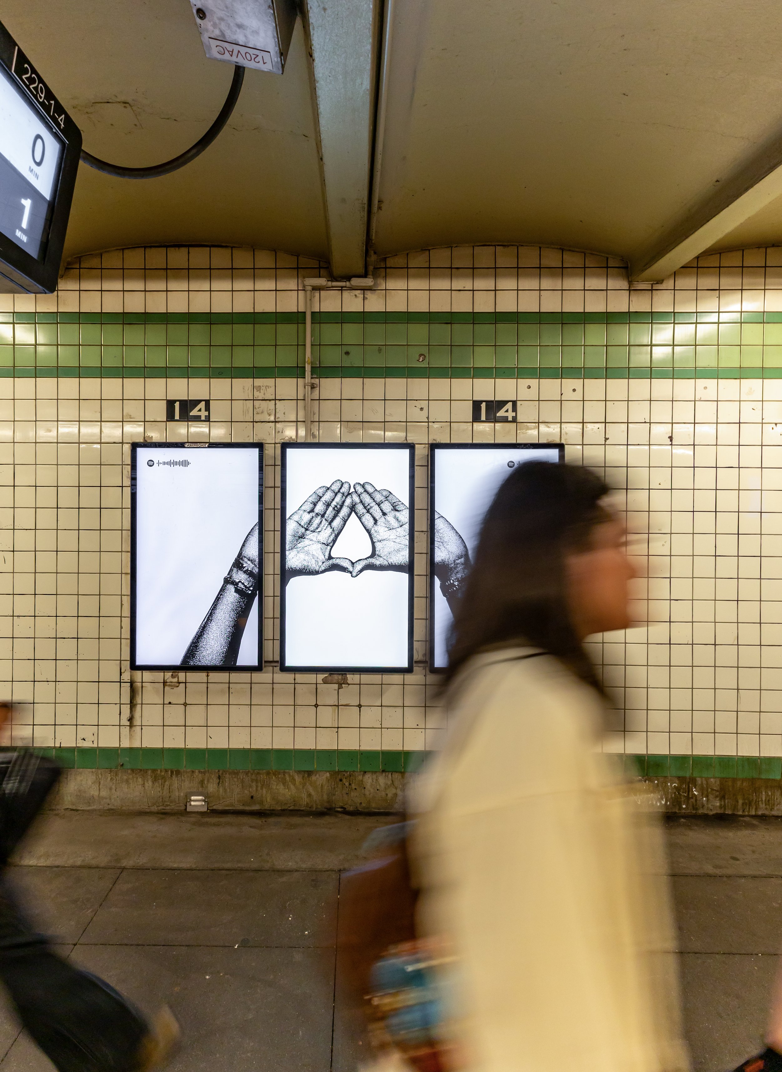 A person walking past three digital advertisements on a tiled wall in a subway station, with one image showing hands forming a heart shape for Jaÿ-Z.