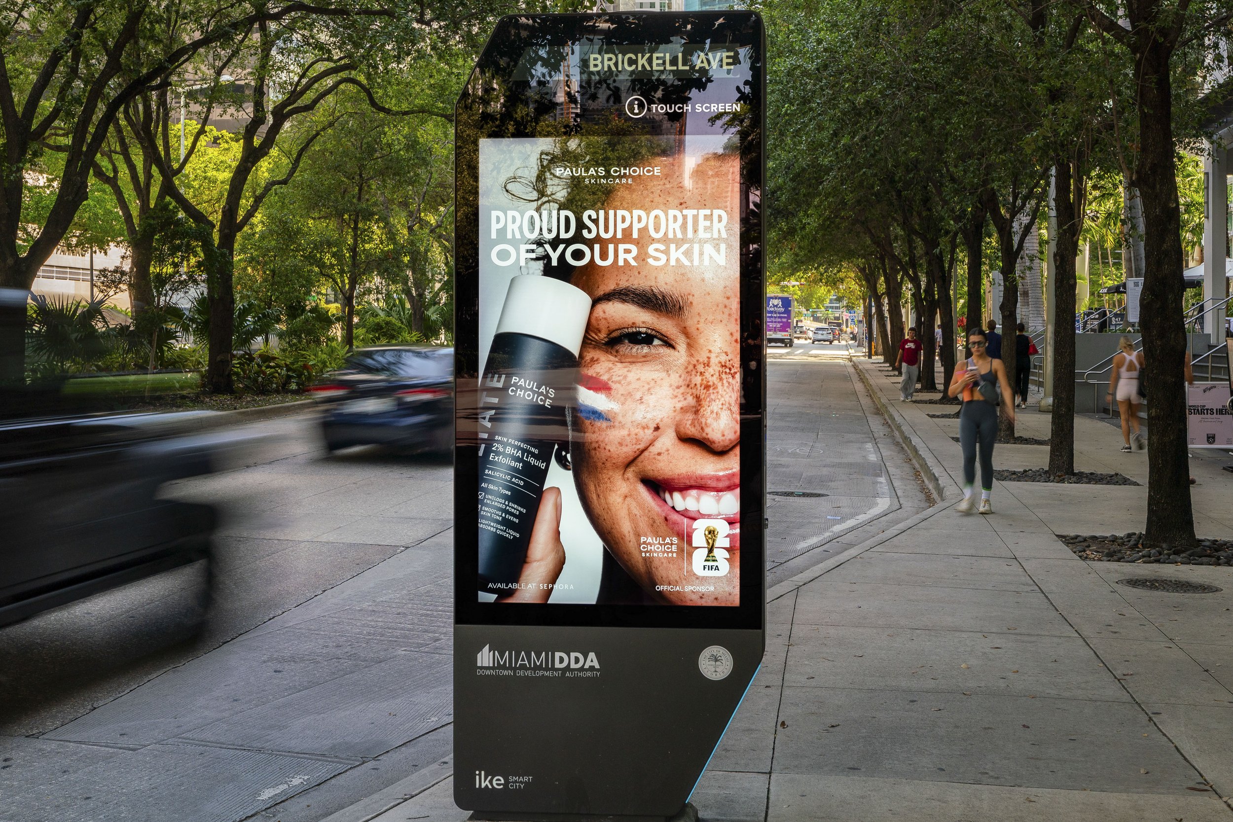 Digital advertising display on a sidewalk with an image of a smiling woman with freckles, promoting skincare products. The display includes the text "PROUD SUPPORTER OF YOUR SKIN" and mentions Paula's Choice Skincare, FIFA sponsorship, and availabili