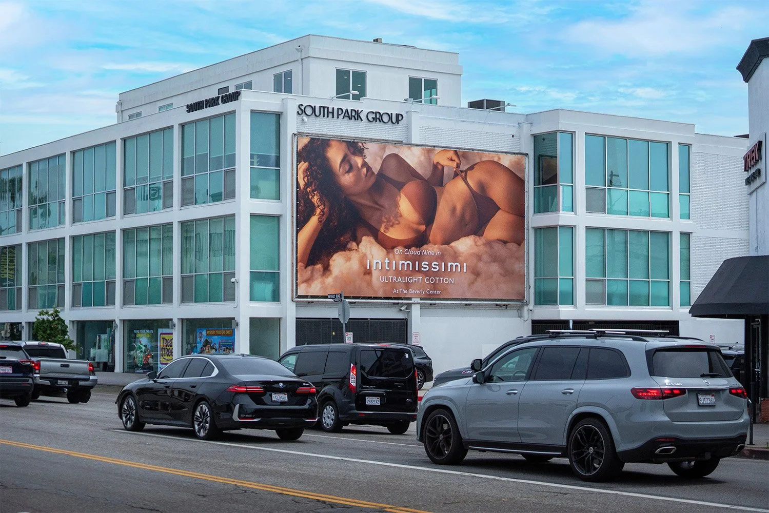 Billboard advertisement on a building for lingerie brand Intimissimi, featuring a woman in black lingerie lying on pink clouds, with the text 'On Cloud Nine in Intimissimi UltraLight Cotton at The Beverly Center.' Several parked cars are on the stree