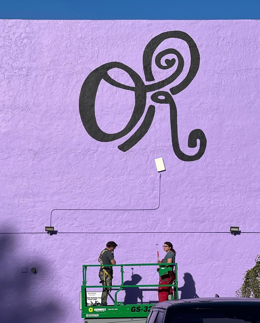 Two people working on a large purple wall with a black artistic design. They are standing on a green lift with shadows cast on the wall.