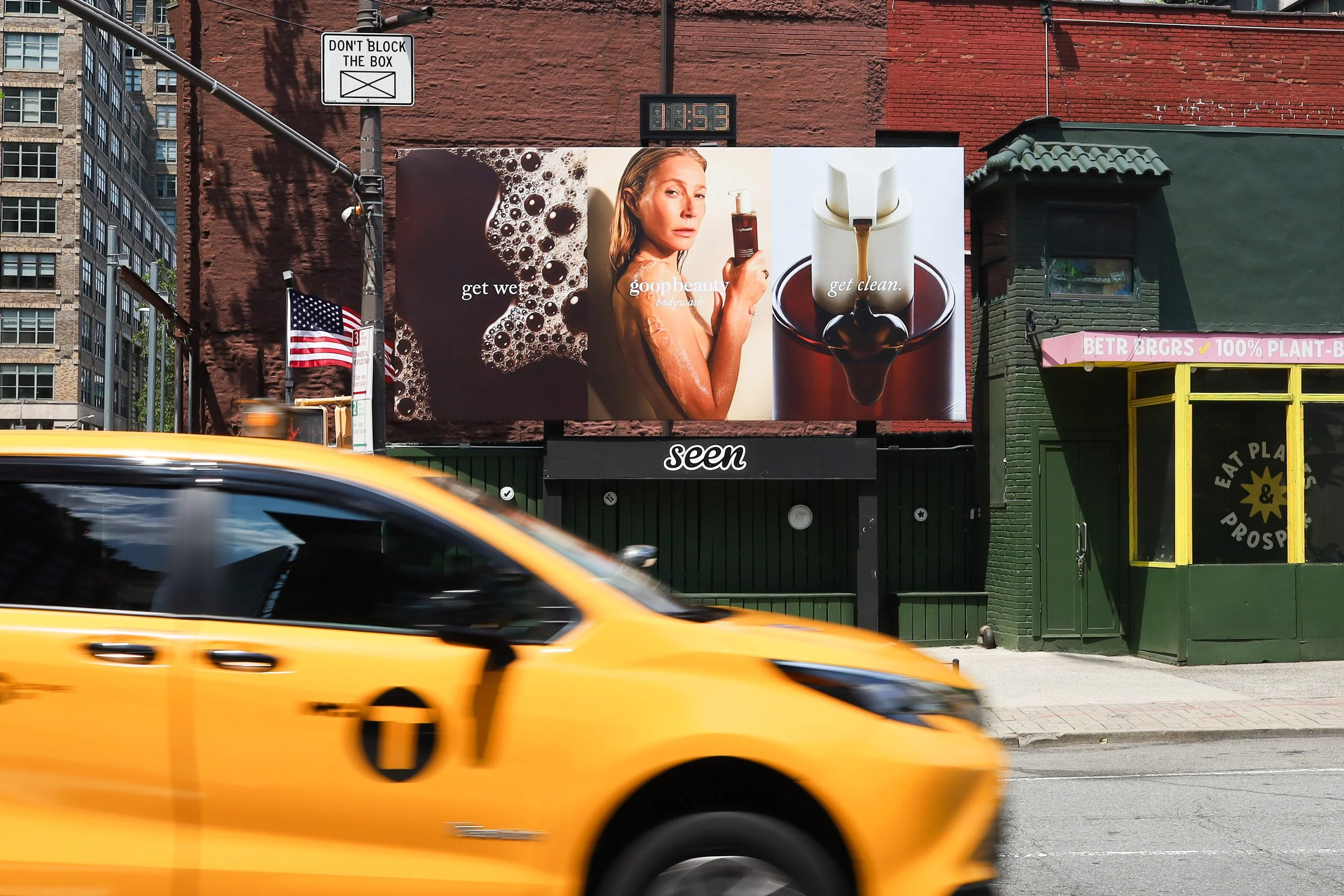 A yellow taxi cab in motion on a city street with buildings, billboards, and traffic signs in the background. The billboard features an advertisement with a woman holding a body wash bottle, soap bubbles, and a soap dispenser, with text "get wet," "g