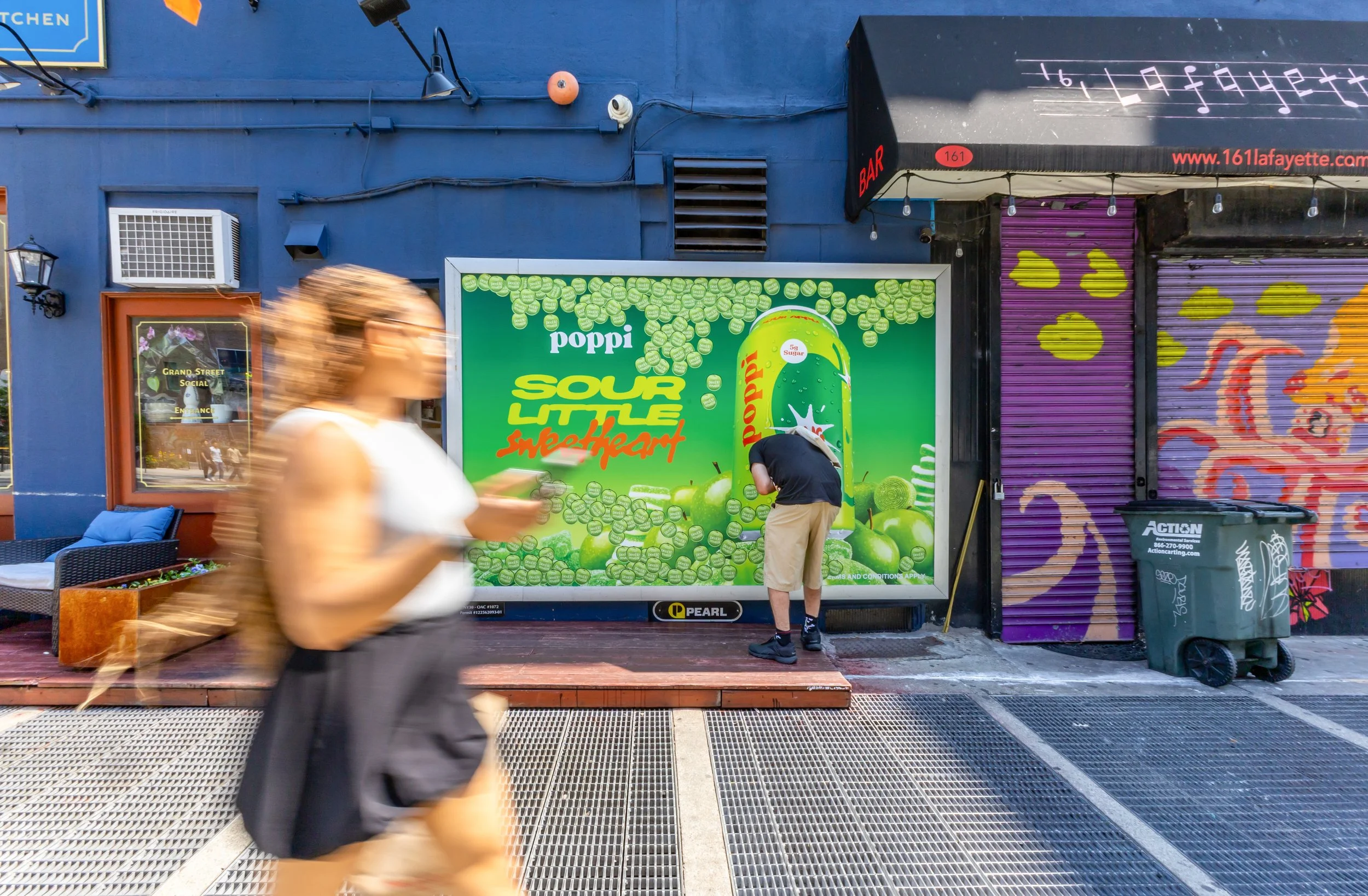 A colorful urban scene with a blue building, a green billboard advertising Poppi sour apple soda, and a purple shutter with yellow graffiti. Two people are walking and one person is bending down near the billboard, with motion blur indicating movement.