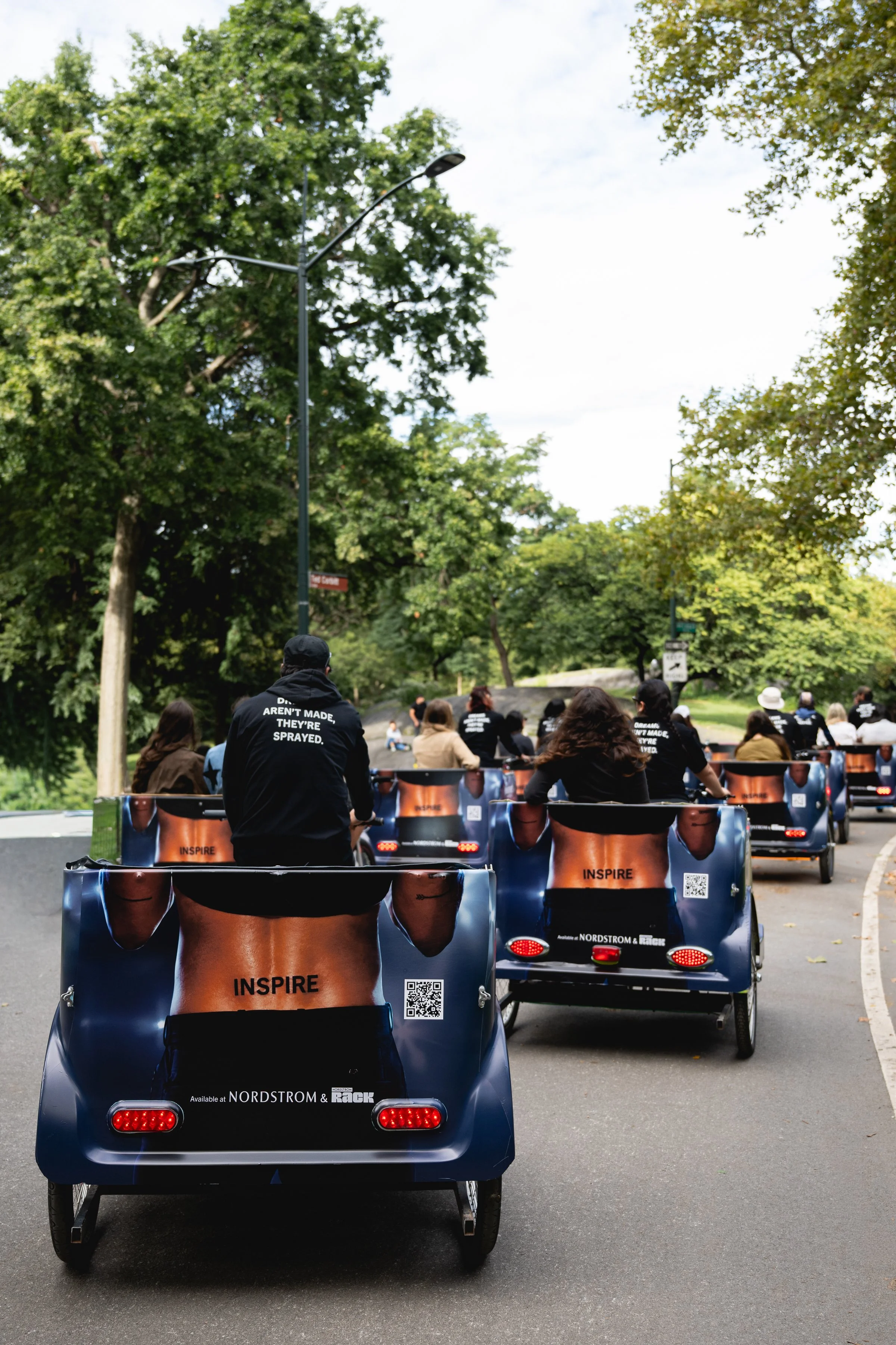 People riding pedal-powered carts on a paved road in a park, surrounded by green trees on a cloudy day.