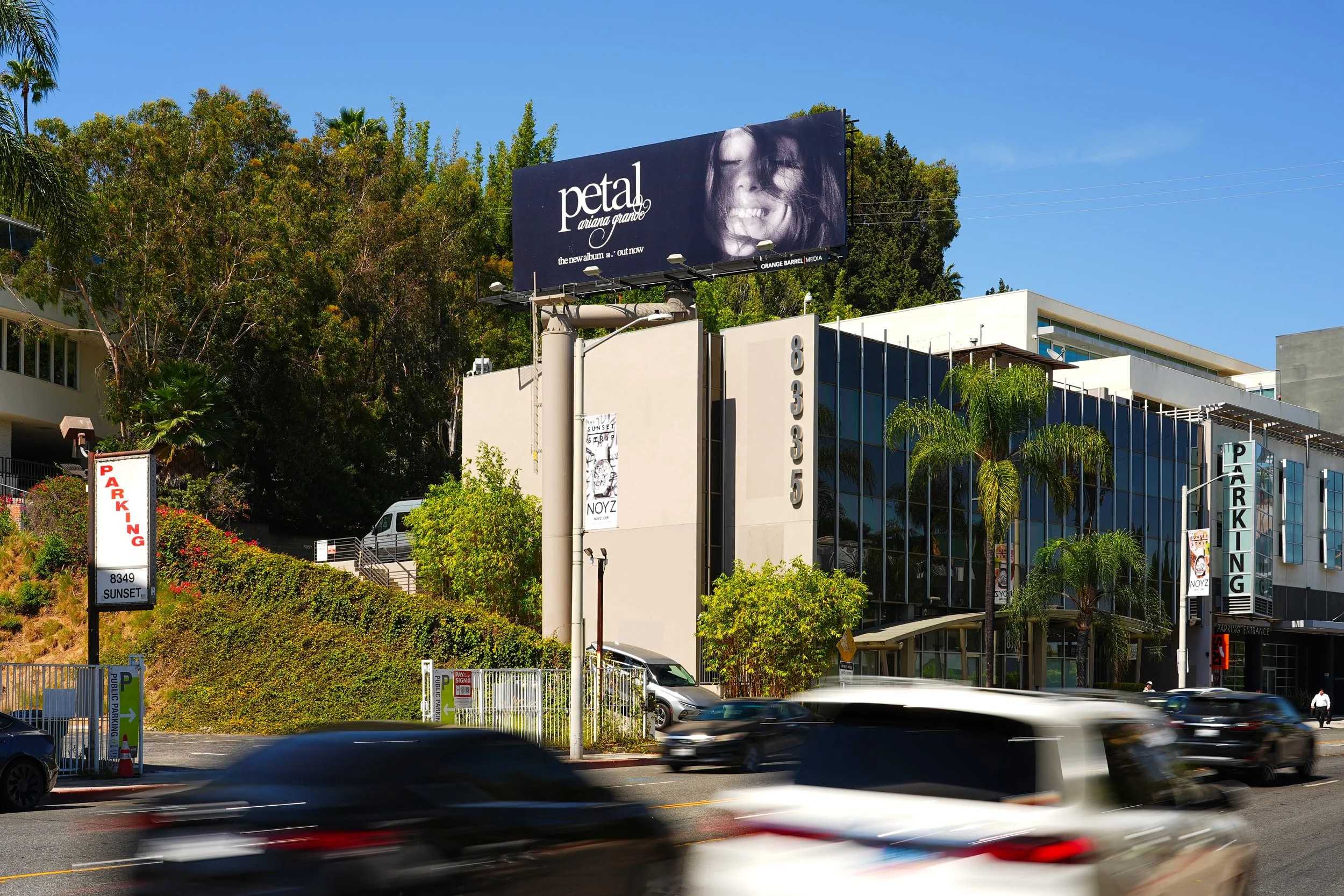 Billboard advertising Ariana Grande's new album titled 'petal,' with a photo of her smiling face and her name in white text on a dark background. Surrounding buildings and moving cars are also visible.