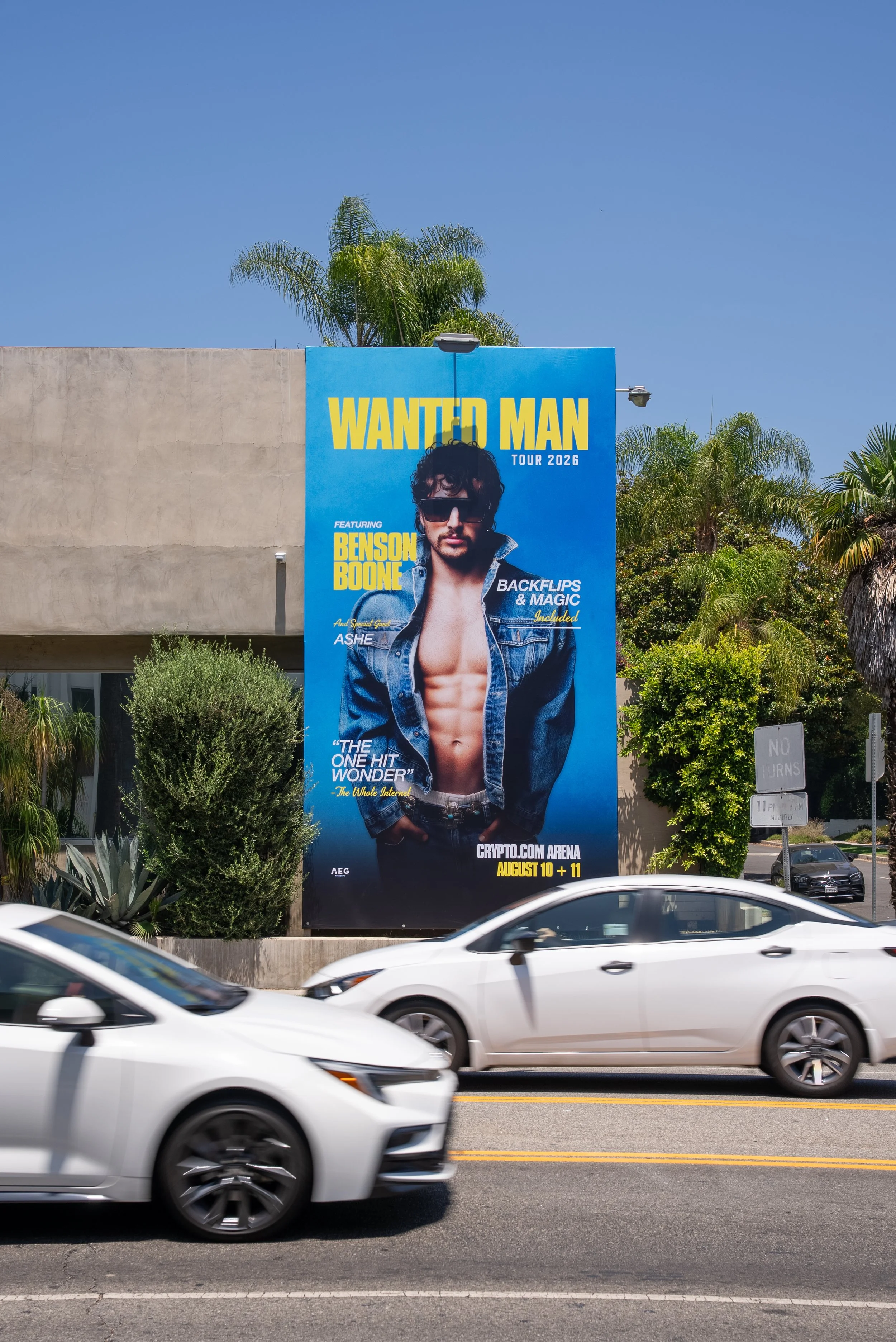 Large blue billboard advertising a music tour featuring Benson Boone at Crypto.com Arena on August 10 and 11, with an image of Benson Boone wearing sunglasses and open denim jacket, showing his chest and abs, with trees and a concrete building in the