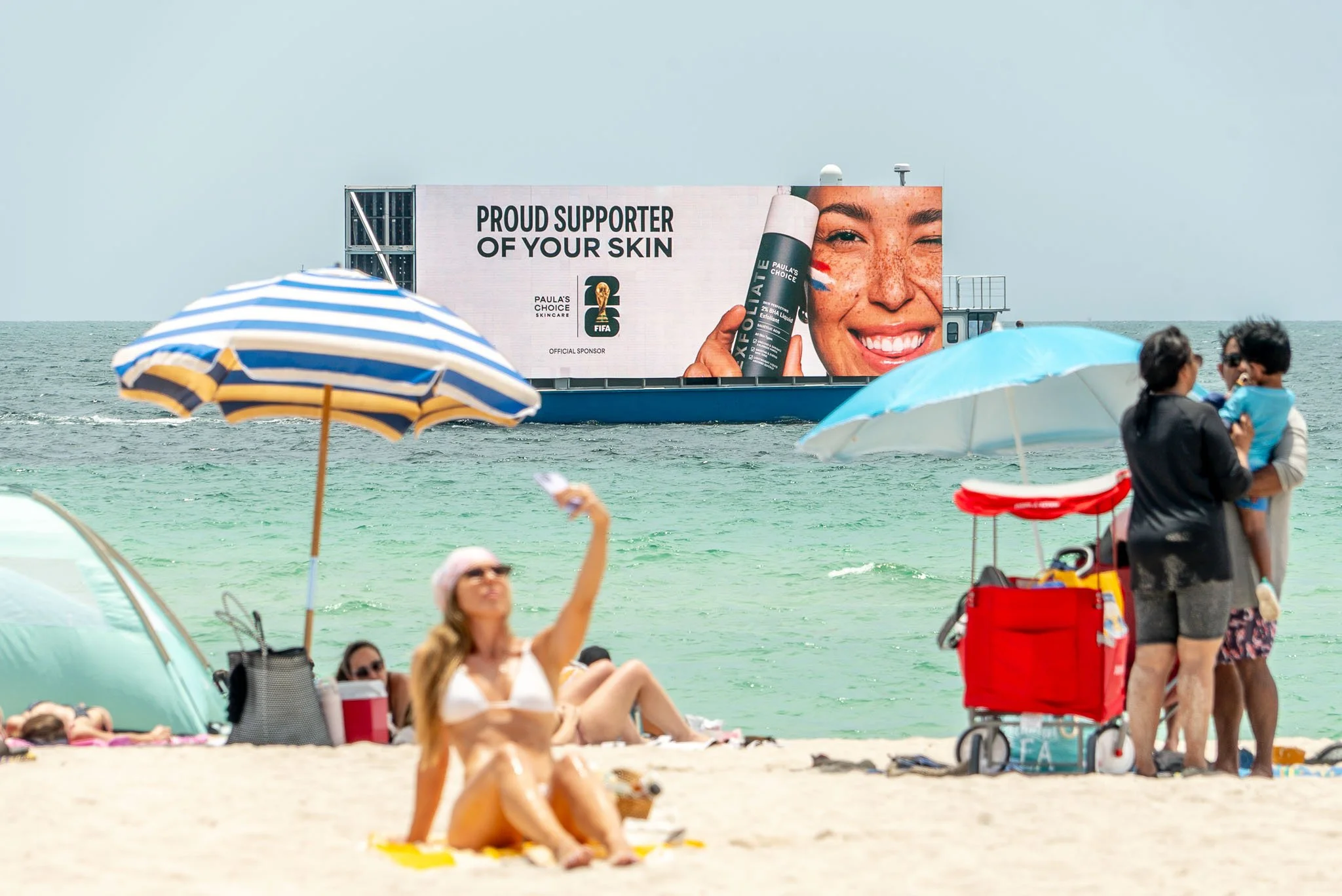 A busy beach scene with people under umbrellas and on the sand, with a large billboard in the background advertising skincare products.