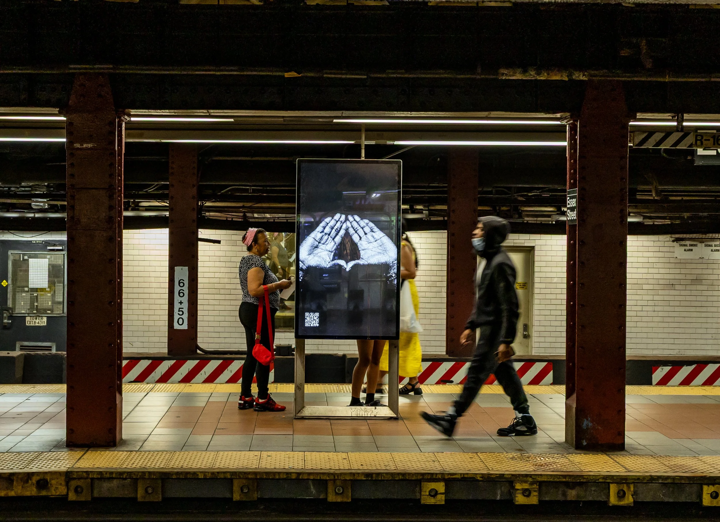 People standing and walking on a subway platform with a digital advertisement display in the center and exposed brick wall in the background.