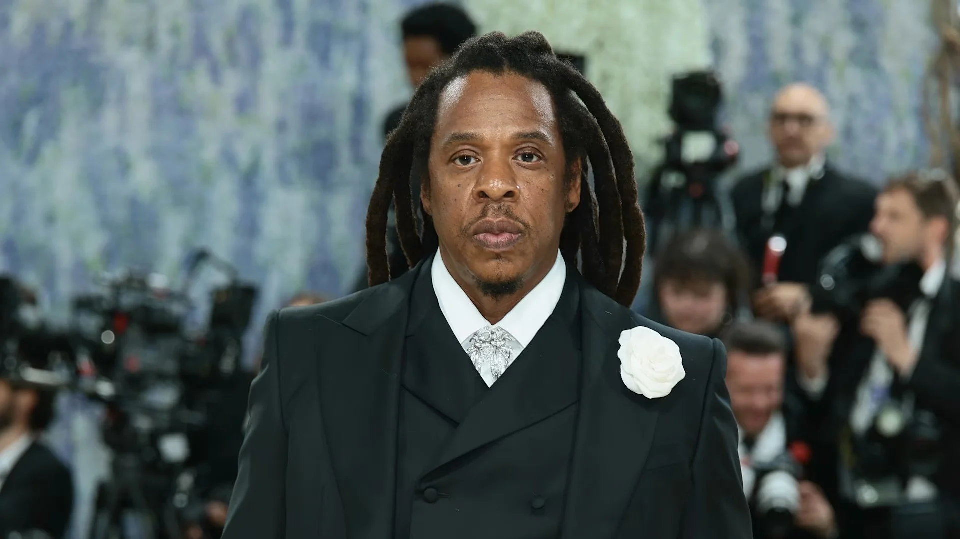 Jaÿ-Z with long dreadlocks wearing a black tuxedo with a white boutonniere, standing in a formal setting with photographers and people in tuxedos in the background.