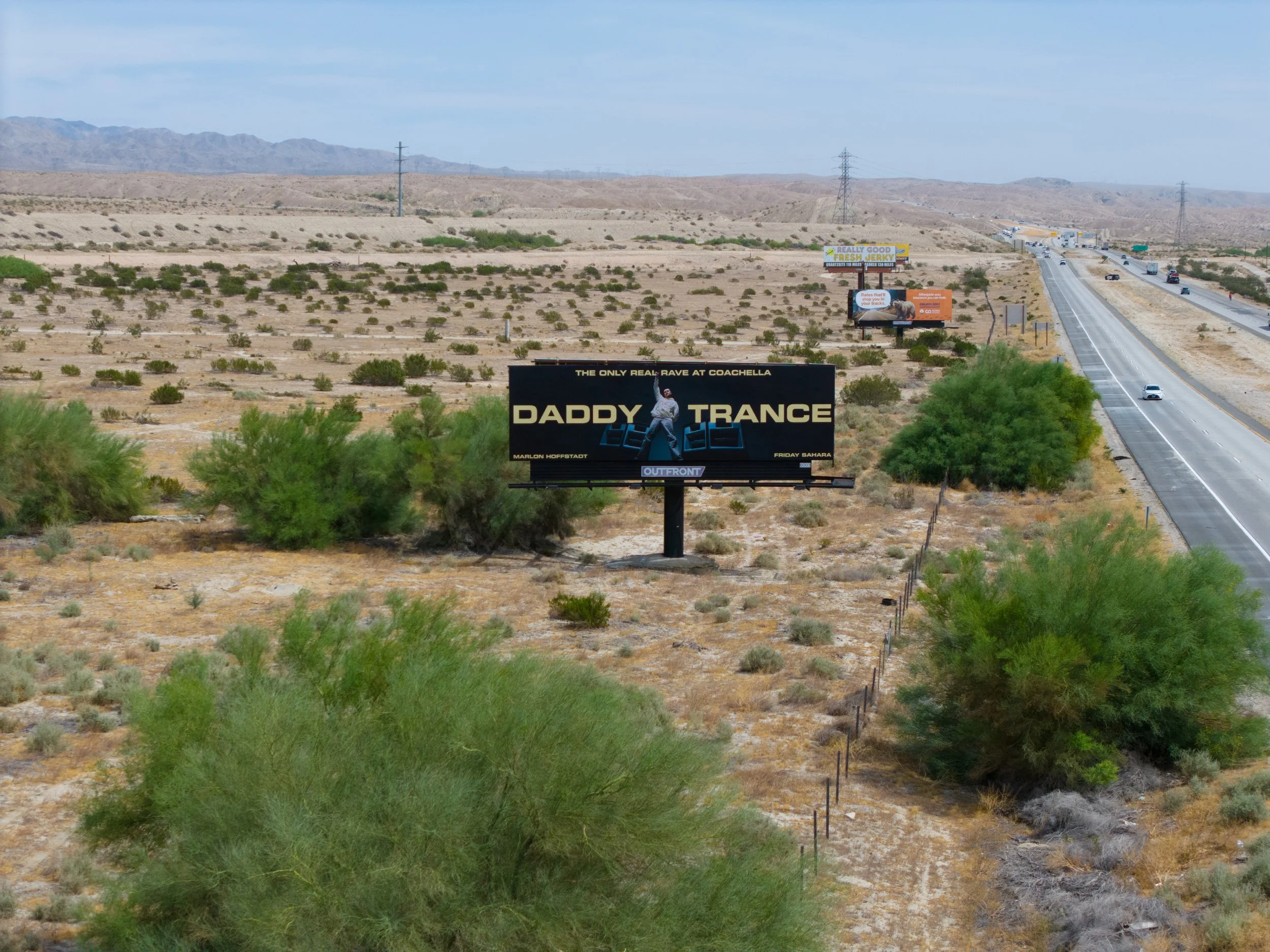 Billboard in a desert landscape advertising Daddy Trance, a rave at Coachella. The billboard features a person with raised arm and speakers, with text indicating it's the only real rave at Coachella, scheduled for Friday Sahara.