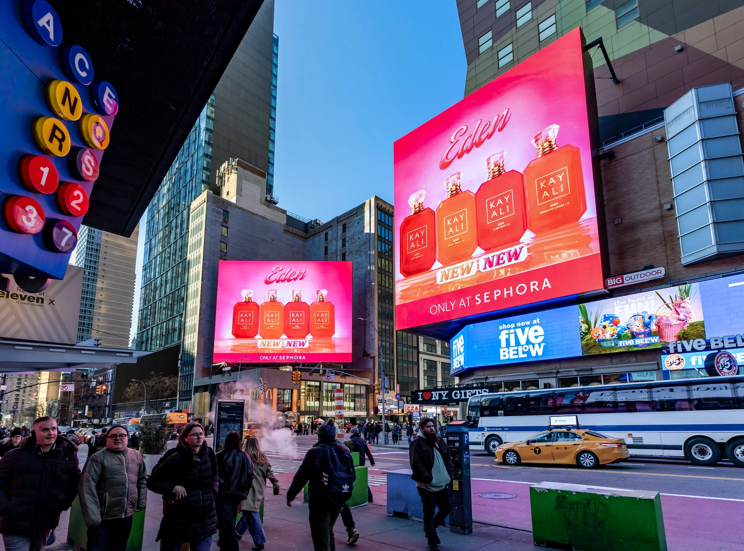 Urban street scene in Times Square, New York City, with people walking, digital billboards advertising Kayali perfume, and city vehicles including a yellow taxi and a bus.