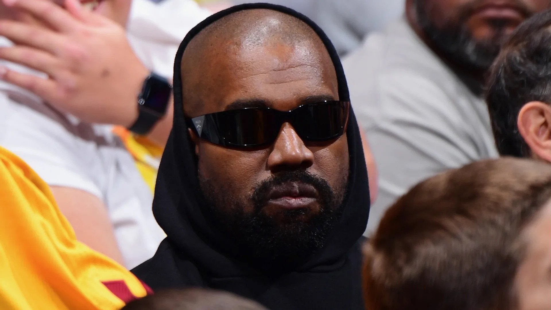 Ye wearing dark sunglasses and a black hoodie, with a beard, seated among others, at an indoor event.