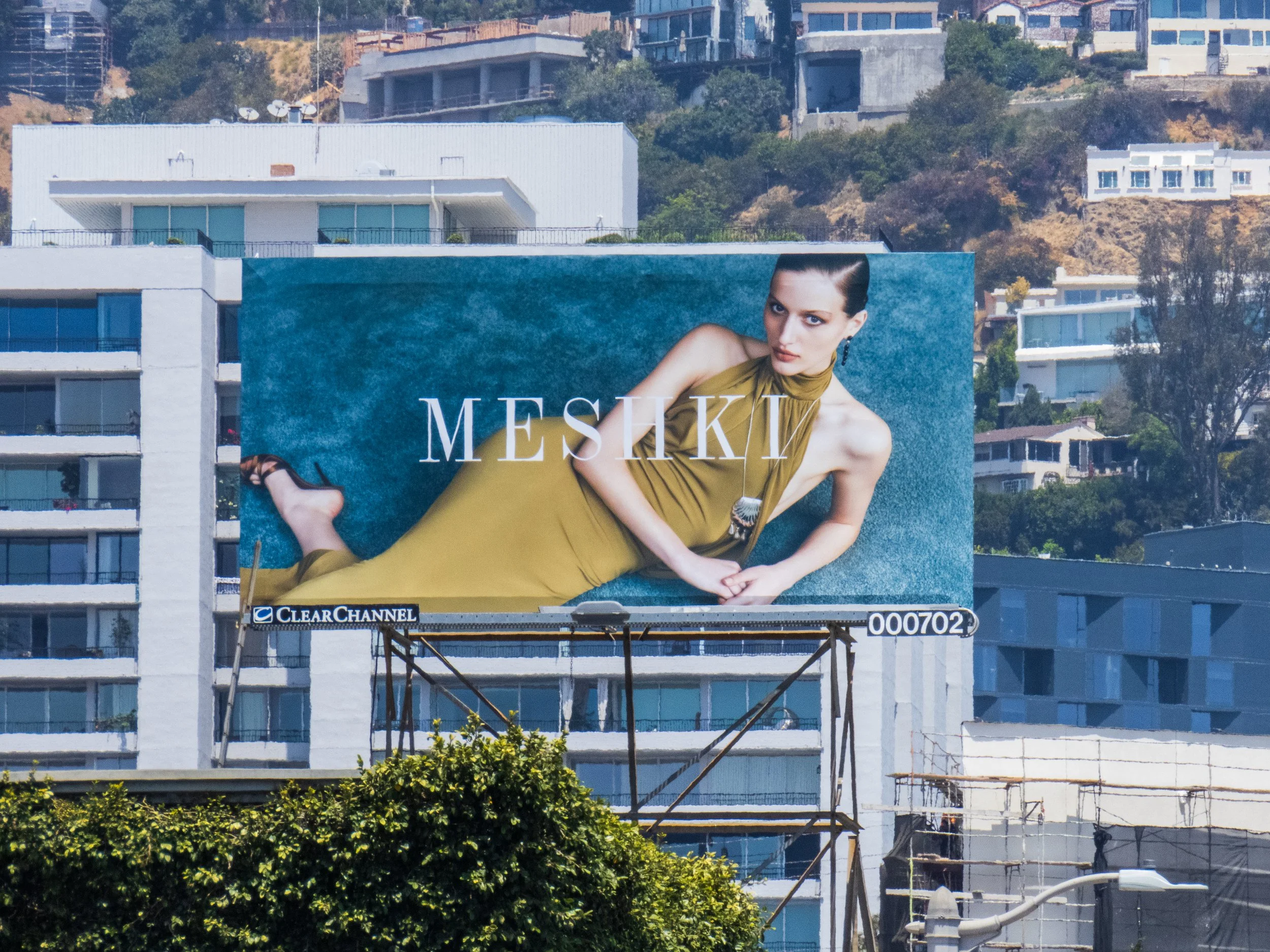 A large billboard featuring a woman in a mustard yellow dress with a serious expression, set against the backdrop of residential buildings and a hillside.