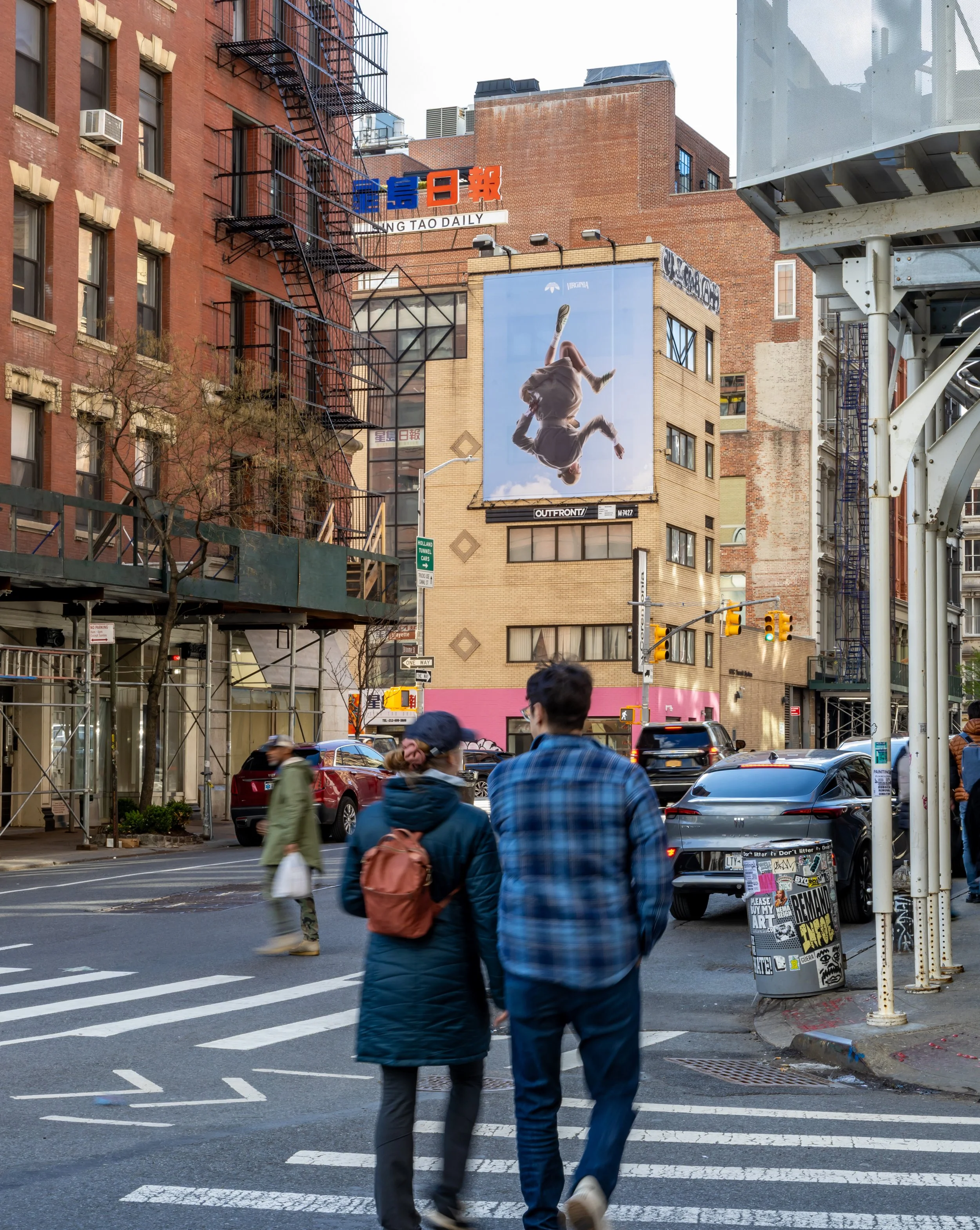 People crossing the street at a city intersection with brick buildings, billboards, and street signs in the background.