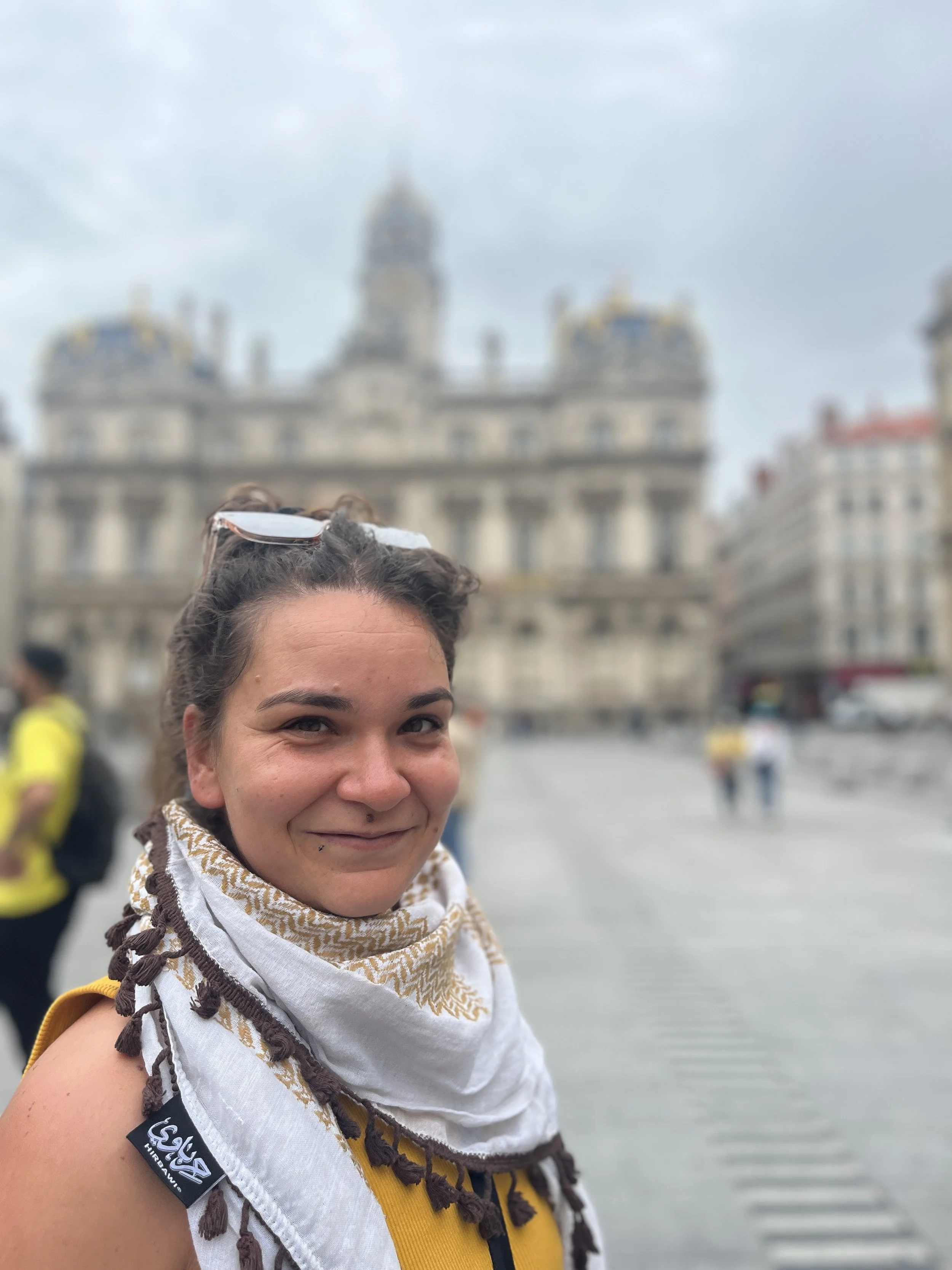 A smiling woman with dark hair wearing a patterned scarf and a yellow backpack standing in front of a historic European building with ornate architecture and a clock tower in the background.