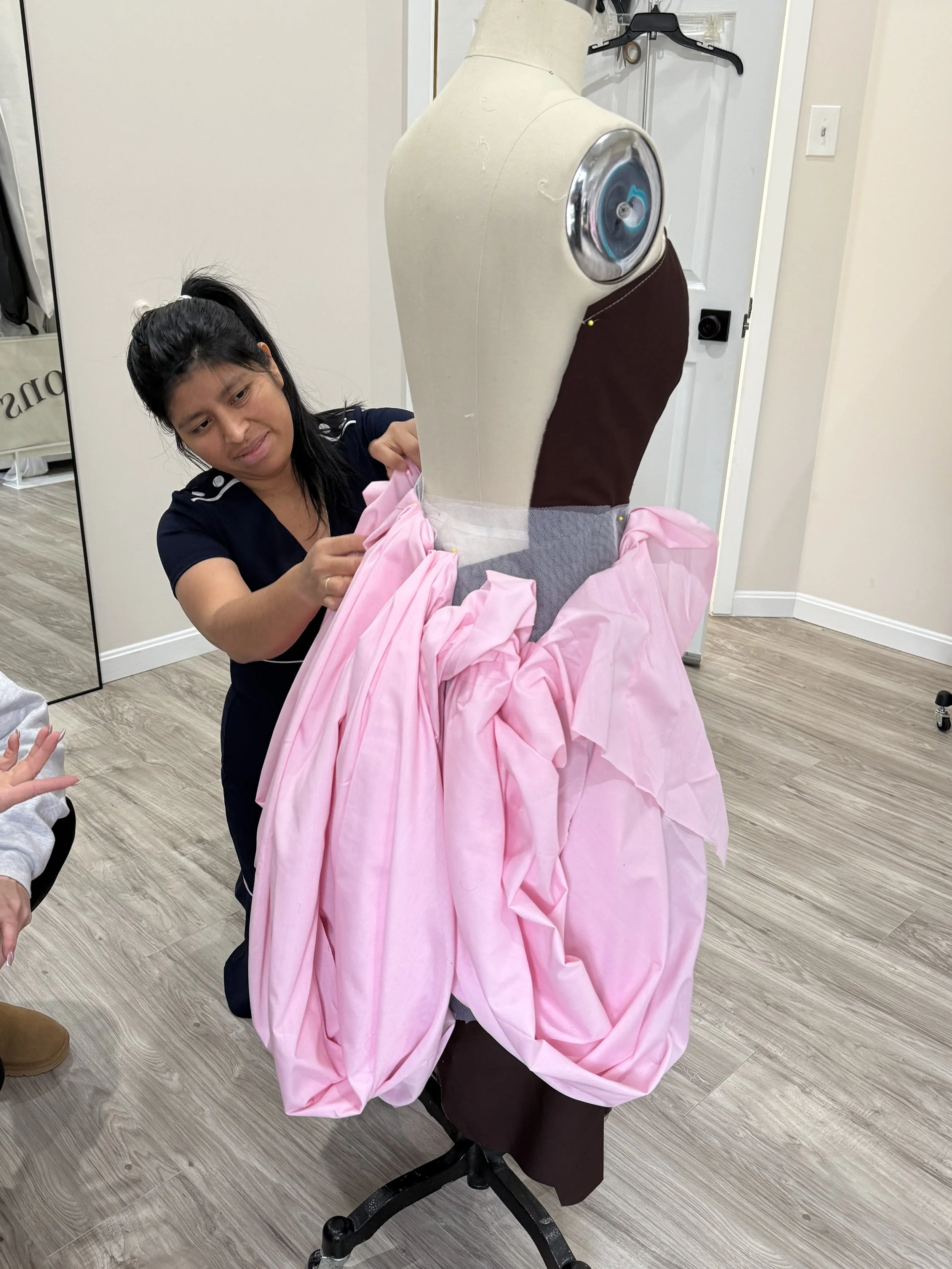 A mannequin dressed in pink fabric and brown clothing, with a metal shoulder fitting, is being adjusted by a woman with dark hair in a ponytail, inside a room with wooden flooring, a mirror, and a white door.