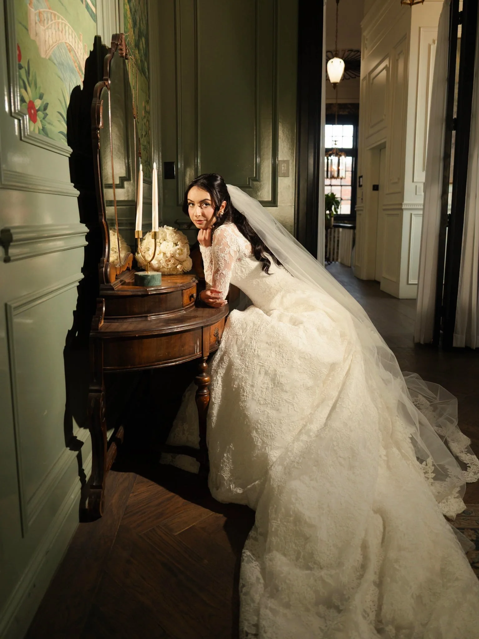 A bride in a white lace wedding dress with a long train and veil is leaning on a vintage wooden dresser with a mirror, flowers, and candles in a room with green walls and wood flooring.