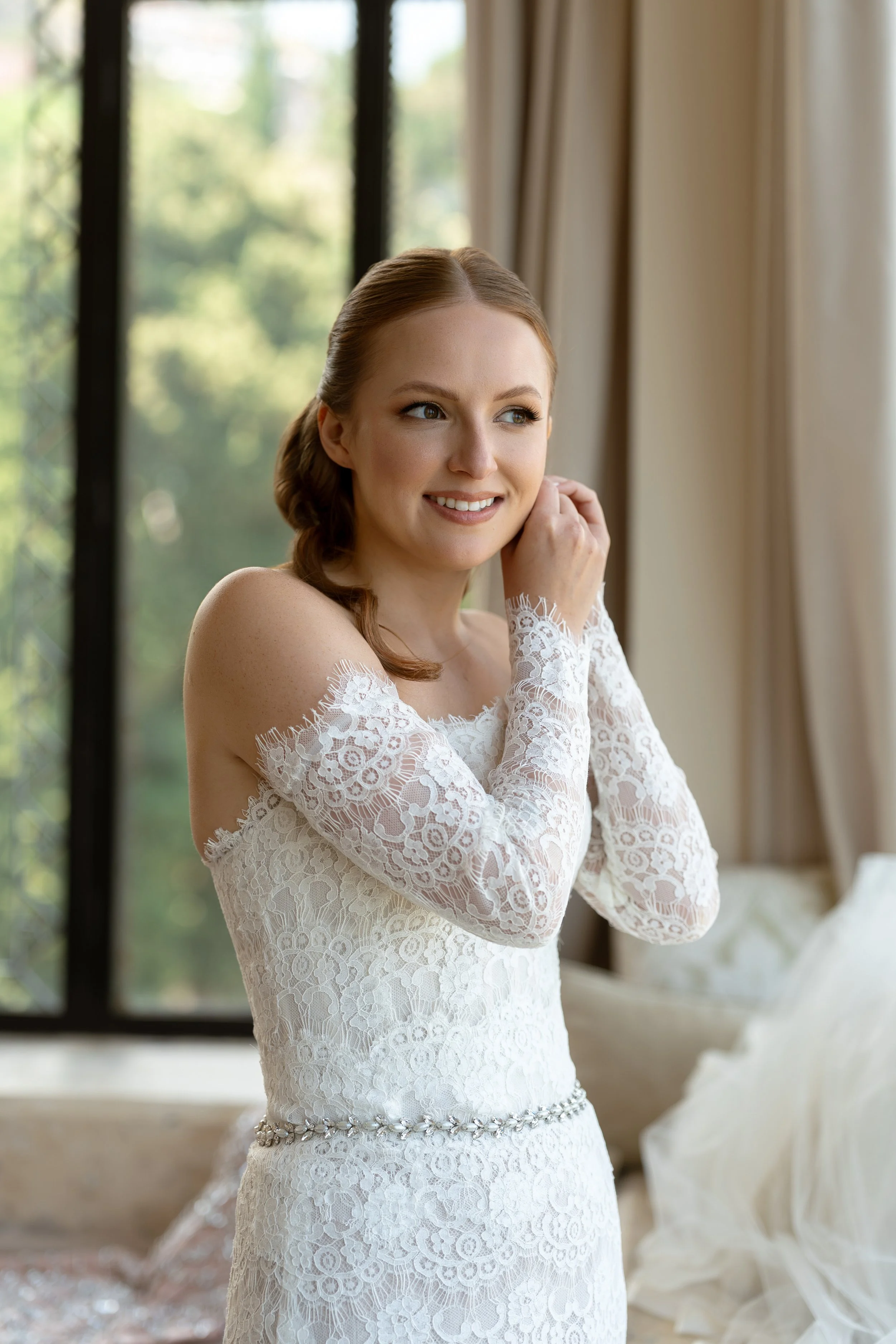 A bride with reddish hair styled in a side braid is smiling and touching her neck. She is wearing a long-sleeve, white lace wedding dress with a pearl belt. She is indoors near large windows with green trees outside, and beige curtains are partially visible.