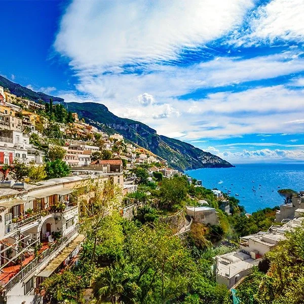 Scenic view of Positano, Italy with colorful hillside buildings, lush greenery, and the Mediterranean Sea under a vibrant blue sky with clouds.
