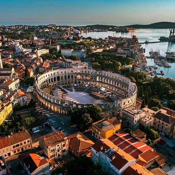 Aerial view of the Pula Arena, an ancient Roman amphitheater in Pula, Croatia, surrounded by urban buildings and near a harbor with boats.