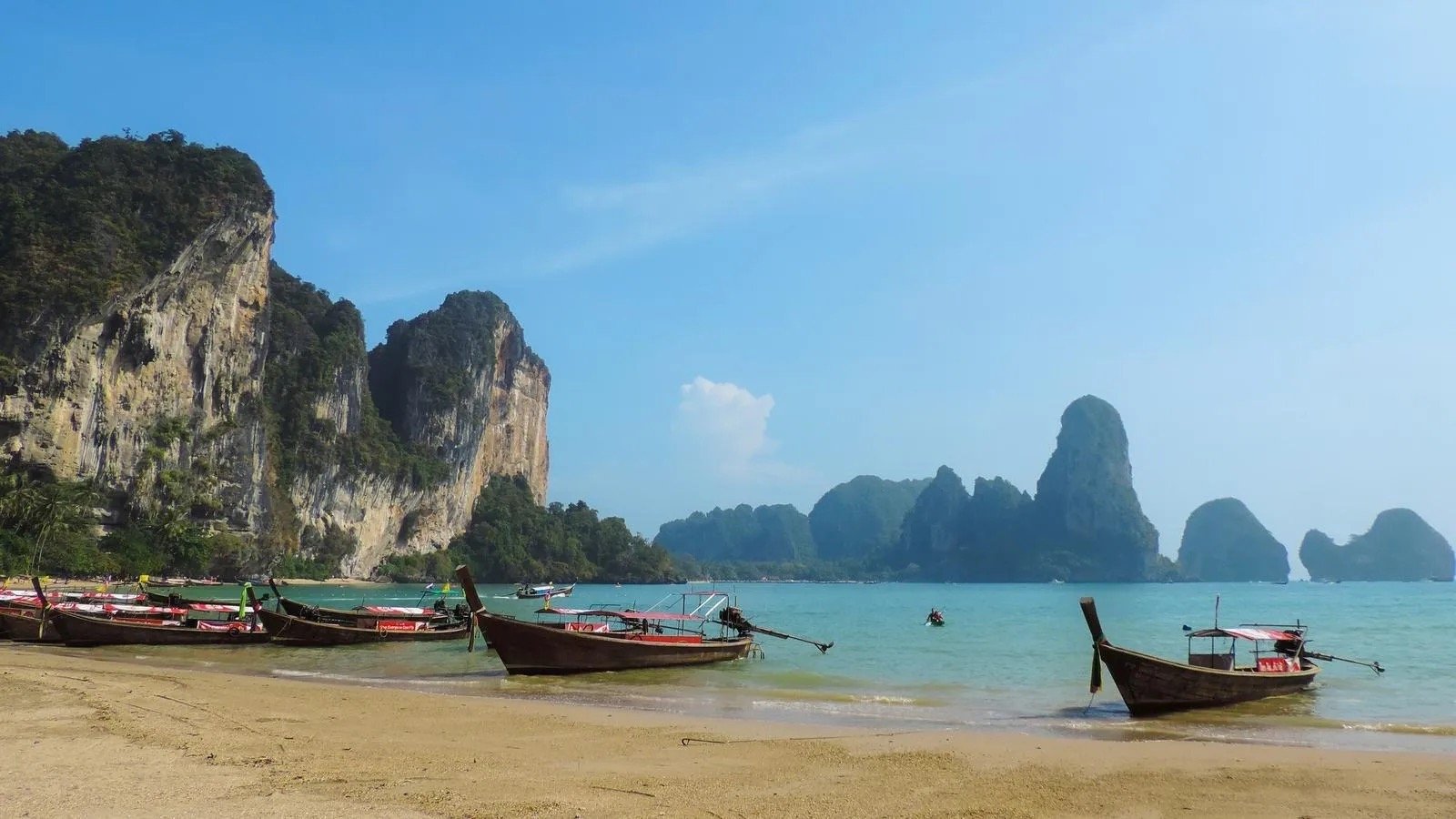 Scenic photo of traditional longtail boats on a sandy beach with turquoise water and dramatic limestone cliffs. Visit Phnom Penh and Angkor Wat, and relax on Thailand's islands.