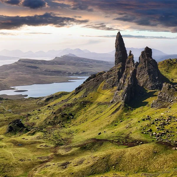A dramatic landscape on the Isle of Skye, Scotland, featuring the iconic jagged rock formations of the Old Man of Storr rising from rolling green hills. The foreground is covered in lush grass and scattered rocks, while a serene loch.