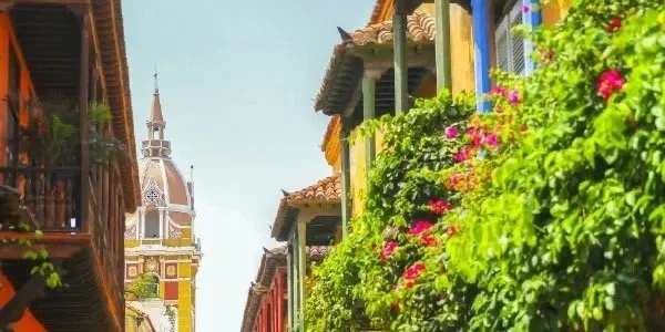 Historic street with colorful buildings, lush greenery, and a church tower in the background.