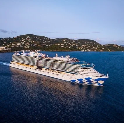 A large white cruise ship with blue wave designs on its bow sails near the coast of St. Thomas, US Virgin Islands. The ship is surrounded by calm blue waters, with lush green hills and scattered houses visible in the background under a clear sky.