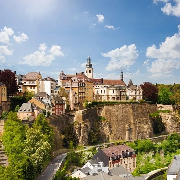 Luxembourg City skyline with historic buildings and fortifications on a cliff; clear blue sky and green foliage.