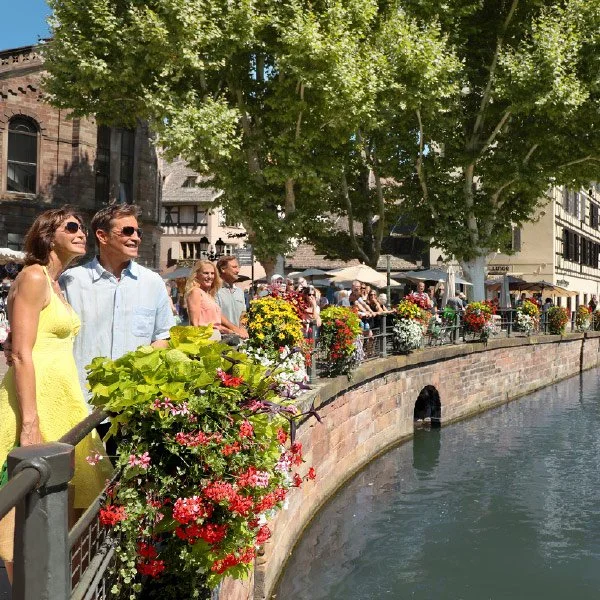 A cheerful couple stands on a flower-adorned bridge over a canal in Strasbourg, France. The scene is lively, with other people enjoying the sunny day along the water. Brightly colored flowers line the bridge, and leafy green trees provide shade.