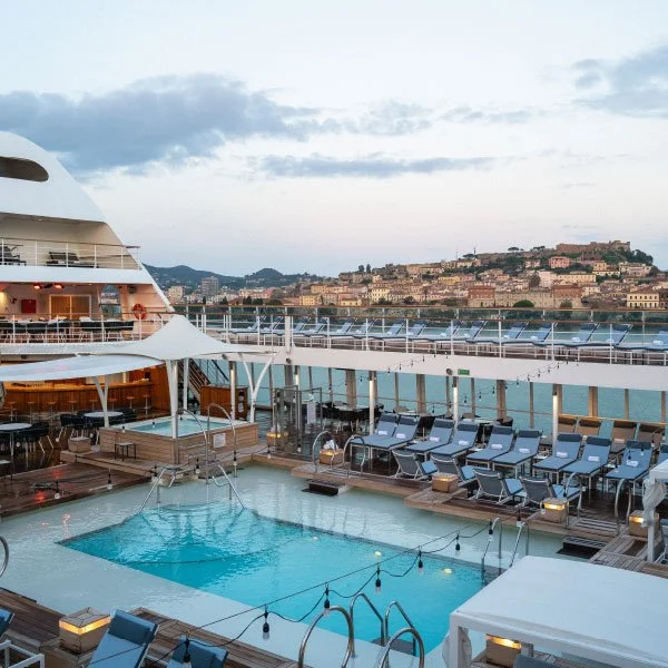 Cruise ship deck with pool and lounge chairs at sunset.