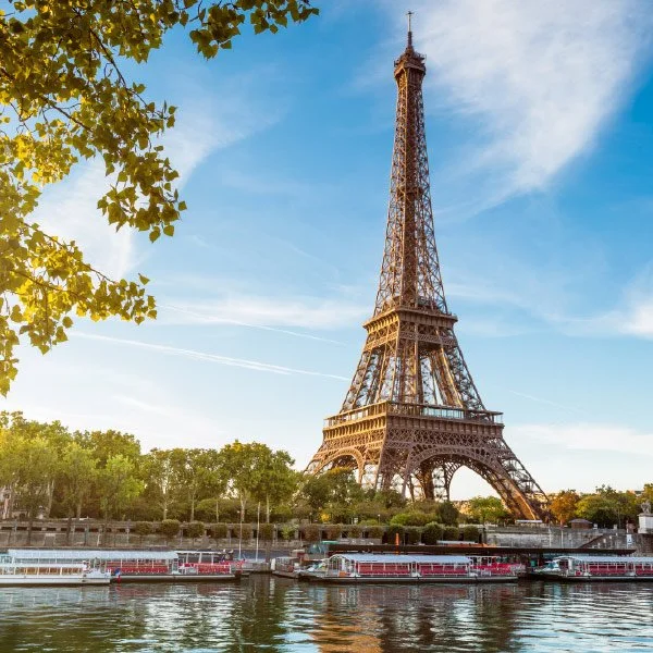 A clear daytime photo of the Eiffel Tower in Paris, France. The iconic iron lattice tower stands tall against a blue sky with wispy clouds. The base of the tower is surrounded by green trees and a river in the foreground, where several boats are dock
