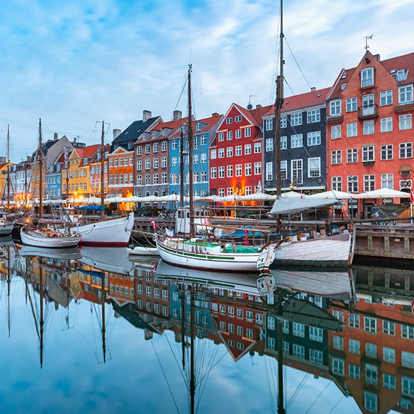 Colorful historic buildings line the Nyhavn waterfront in Copengagen, Denmark, with sailboats docked along the canal and their reflections mirrored in the calm water below under a partly cloudy sky.