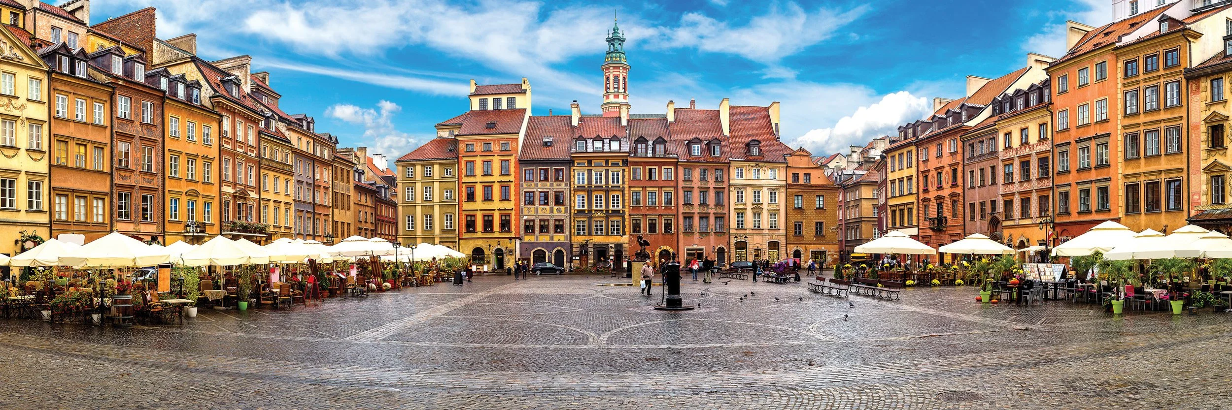 Colorful historic buildings and a central clock tower in Warsaw's Old Town Square, with a cobblestone plaza in the foreground and a few people walking under a blue sky.