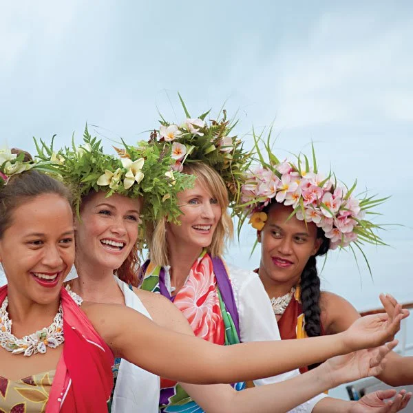Four women wearing floral crowns and colorful traditional Polynesian attire, performing a dance outdoors.