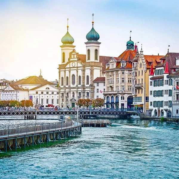 Scenic view of traditional European buildings and a church with two green domes by a river, under a clear blue sky.