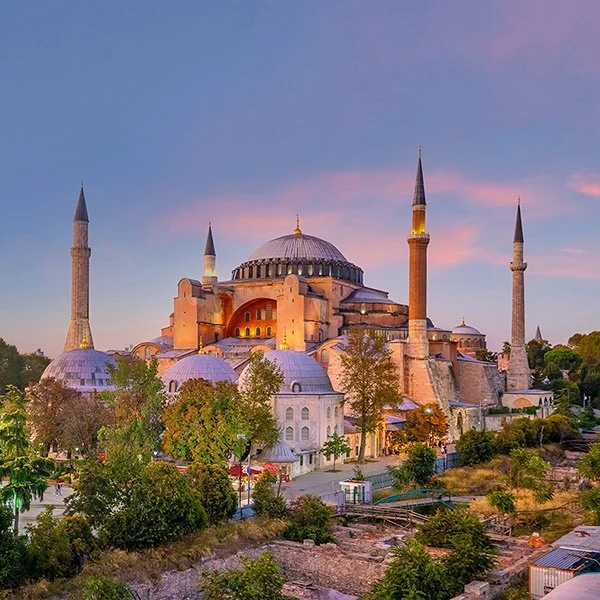 Hagia Sophia in Istanbul at sunset, illuminated by golden lights, showcasing its massive central dome, four slender minarets, and a blend of Byzantine and Ottoman architectural features, surrounded by lush greenery and historic ruins under a pastel