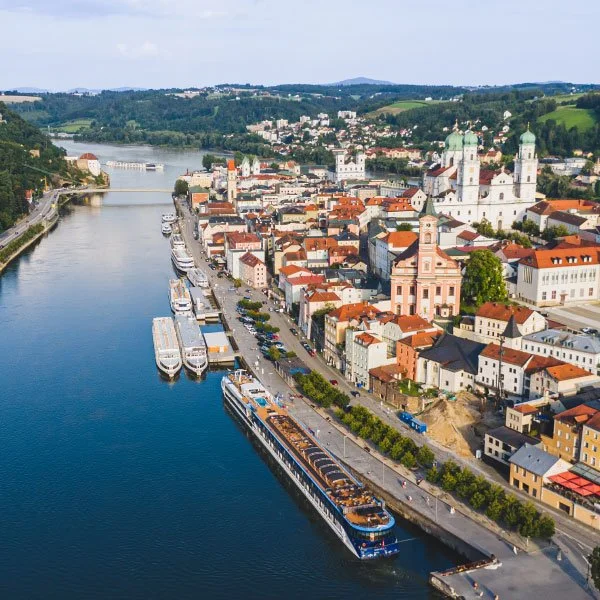 Aerial view of the AmaViola in Passaud, Germanywith a river, boats docked along the shoreline, historic buildings with red roofs, and a church with green domes, surrounded by hills.