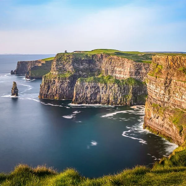 A scenic view of the Cliffs of Moher in Ireland, showing dramatic, steep cliffs rising above the Atlantic Ocean with lush green grass on top and sunlight illuminating the rugged rock faces. The sea below is calm with gentle waves.