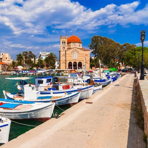 A Greek harbor scene with traditional fishing boats moored alongside a dock. In the background, there's a historic church with a dome and a tower, surrounded by trees and buildings under a partly cloudy sky.