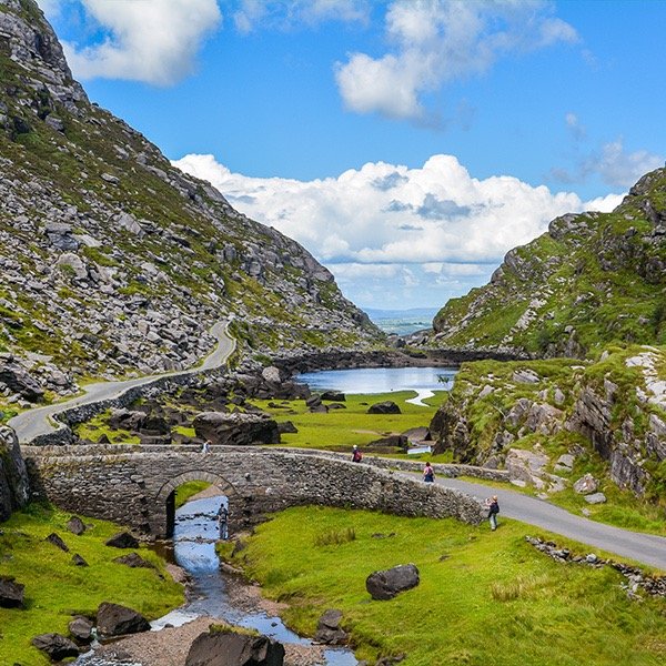 Scenic view of a stone bridge and winding road in a lush green valley with rocky hills and a small stream under a blue sky with clouds.