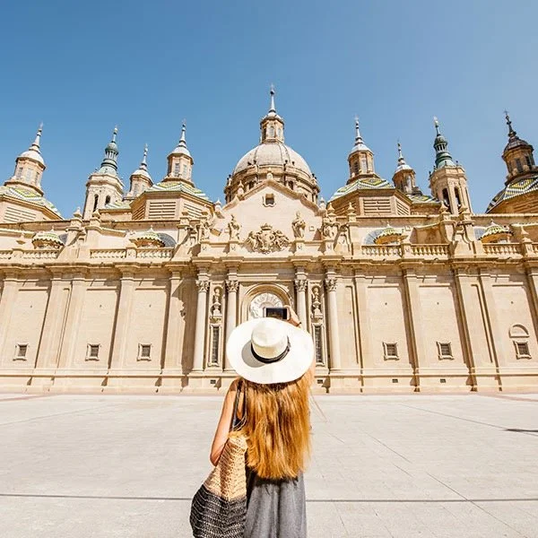A woman with long hair wearing a white hat and a dress stands in front of a large, historical cathedral with intricate architecture and multiple domes under a clear blue sky.