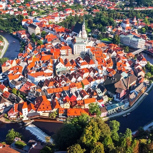 Aerial view of a historic European town with red roofs and a river forming a peninsula. A prominent church with a tall steeple is in the center, surrounded by tightly packed colorful buildings. The landscape is lush with greenery.